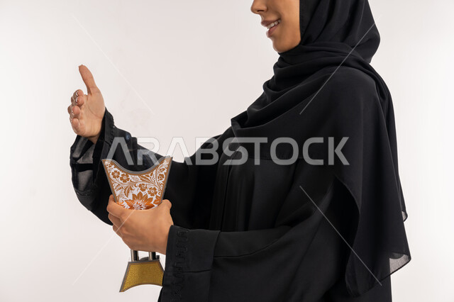 استخدام خشب العود للتطيب بالأعياد و المناسبات, بورتريه مقرب من الجانب لامرأة مبتسمة عربية خليجية إماراتية محجبة ترتدي العباءة السوداء تمسك المبخرة التراثية المزخرفة بيدها, شابة سعودية تقوم بالتعطر بالبخور, خلفية بيضاء