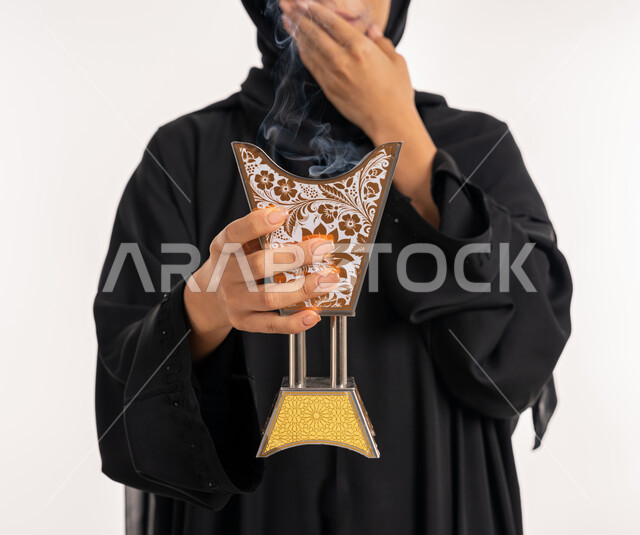 استخدام خشب العود بالأعياد والمناسبات, أصالة العادات والتقاليد الخليجية, بورتريه مقرب لامرأة عربية خليجية إماراتية محجبة ترتدي العباءة السوداء تمسك بيدها المبخرة التراثية المزخرفة, شابة سعودية تقوم بالتعطر بالبخور, خلفية بيضاء