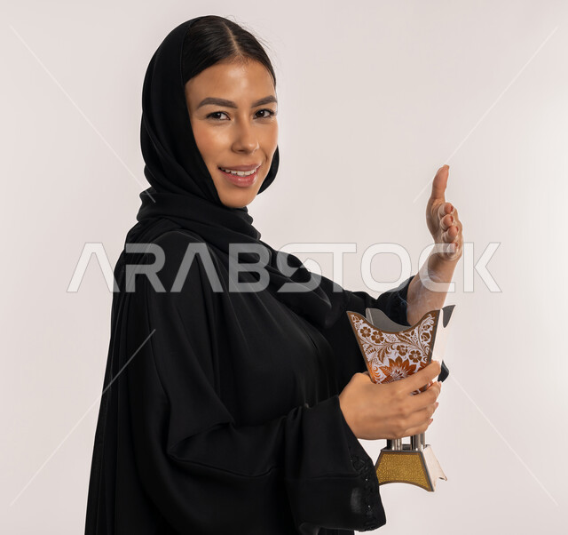 The authenticity of Gulf customs and traditions, the use of agarwood on holidays and occasions, a close-up portrait of a veiled Arab Gulf Emirati woman wearing a black abaya, holding the decorated traditional incense burner in her hand and looking at the camera with gestures of happiness and pleasure, a young Saudi woman perfuming herself with incense, white background