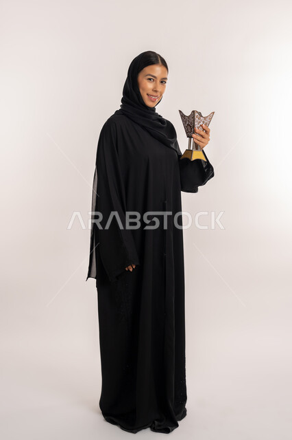 Looking at the camera with gestures of happiness and pleasure, using agarwood for perfume, portrait of a veiled Gulf Arab Emirati woman wearing a black abaya holding an ornate traditional incense burner in her hand, a young Saudi woman perfuming herself with incense, full-length body portrait, white background