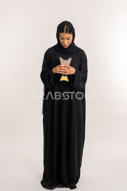 استخدام خشب العود للتطيب, أصالة العادات والتقاليد الخليجية, بورتريه لامرأة عربية خليجية إماراتية محجبة ترتدي العباءة السوداء تمسك المبخرة التراثية المزخرفة بيدها و تنظر اليها, شابة سعودية تقوم بالتعطر من حامل البخور, صورة للجسم بالطول الكامل, خلفية بيضاء