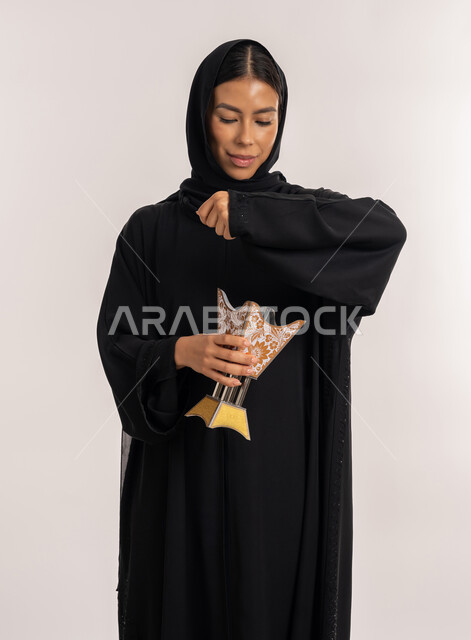 أصالة العادات والتقاليد الخليجية, استخدام خشب العود للتطيب, بورتريه لامرأة عربية خليجية إماراتية محجبة ترتدي العباءة السوداء تمسك المبخرة التراثية المزخرفة بيدها, شابة سعودية تقوم بالتعطر من حامل البخور, منتجات محلية وطنية, خلفية بيضاء