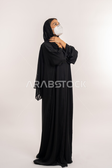 Following precautionary measures to prevent the Corona virus, portrait of a veiled Arab Gulf Emirati woman wearing a black abaya, placing her hands on her neck with suffocation gestures, a young Saudi woman feeling difficulty breathing, full-length body portrait, white background