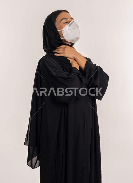 اتباع الإجراءات الاحترازية للوقاية من فايروس كورونا, الحفاظ على السلامة, بورتريه لامرأة عربية خليجية إماراتية محجبة ترتدي العباءة السوداء و الكمامة تضع يديها على عنقها بإيماءات الاختناق, شابة سعودية تشعر بالاختناق, خلفية بيضاء