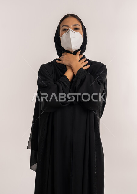 الحفاظ على السلامة, اتباع الإجراءات الاحترازية للوقاية من فايروس كورونا, بورتريه لامرأة عربية خليجية إماراتية محجبة ترتدي العباءة السوداء و الكمامة تضع يديها على عنقها بإيماءات الاختناق, شابة سعودية تشعر بصعوبة بالتنفس, خلفية بيضاء