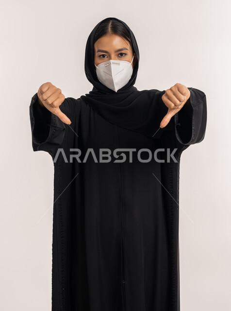 Expressions of disapproval and rejection, avoiding the spread of infection from Covid-19, a portrait of a veiled Arab Gulf Emirati woman wearing a black abaya and a mask, following precautionary measures to prevent the Corona virus, a young Saudi woman pointing with two thumbs down, white background