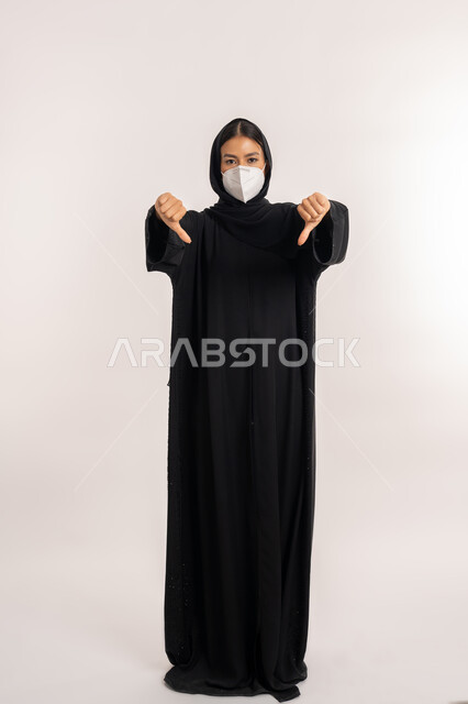 تعابير عدم الموافقة و الرفض ,تجنب انتشار العدوى من كوفيد 19، بورتريه لامرأة عربية خليجية إماراتية محجبة ترتدي العباءة السوداء و الكمامة تتبع الإجراءات الاحترازية للوقاية من فايروس كورونا, شابة سعودية تشير بإصبعي الإبهام للأسفل, صورة للجسم بالطول الكامل, خلفية بيضاء