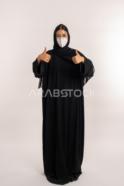 إيماءات الموافقة و القبول, تجنب انتشار العدوى من كوفيد 19، بورتريه لامرأة عربية خليجية إماراتية محجبة ترتدي العباءة السوداء و الكمامة تتبع الإجراءات الاحترازية للوقاية من فايروس كورونا, شابة سعودية تشير بإصبعي الإبهام للأعلى, صورة للجسم بالطول الكامل, خلفية بيضاء