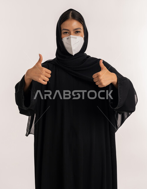 Expressions of approval and acceptance, avoiding the spread of infection from Covid-19, a portrait of a veiled Emirati Gulf Arab woman wearing a black abaya and a mask, following precautionary measures to prevent the Corona virus, a young Saudi woman pointing upward with her thumb, white background