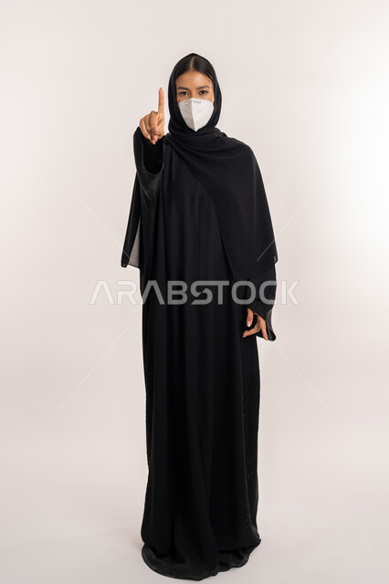 إشارة تمثل الرقم واحد, الحفاظ على المسافات وعدم الملامسة السطحية, بورتريه لامرأة عربية خليجية إماراتية محجبة ترتدي العباءة السوداء و الكمامة تتبع الاحترازات الوقائية ضد كوفيد 19, شابة سعودية تشير بإصبع السبابة إلى الأعلى, صورة للجسم بالطول الكامل, خلفية بيضاء