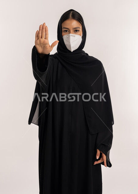 الحفاظ على المسافات وعدم الملامسة السطحية, بورتريه لامرأة عربية خليجية إماراتية محجبة ترتدي العباءة السوداء و الكمامة تتبع الاحترازات الوقائية ضد كوفيد 19, شابة سعودية ترفع يدها بتعابير التوقف و تجنب المصافحة, خلفية بيضاء