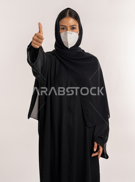 تجنب انتشار العدوى من كوفيد 19، بورتريه لامرأة عربية خليجية إماراتية محجبة ترتدي العباءة السوداء و الكمامة تتبع الإجراءات الاحترازية للوقاية من فايروس كورونا, شابة سعودية تشير بإصبع الإبهام إلى الأعلى بتعابير الموافقة و القبول, خلفية رمادية