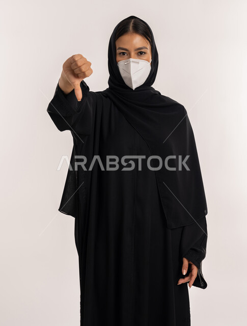 تجنب انتشار العدوى من كوفيد 19, بورتريه لامرأة عربية خليجية إماراتية محجبة ترتدي العباءة السوداء و الكمامة تتبع الإجراءات الاحترازية للوقاية من فايروس كورونا, شابة سعودية تشير بإصبع الإبهام إلى الأسفل بتعابير عدم الإعجاب والموافقة, خلفية بيضاء