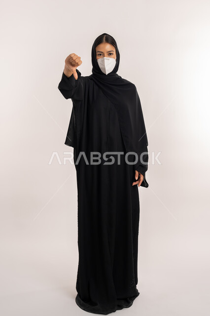 تجنب انتشار العدوى من كوفيد 19, بورتريه لامرأة عربية خليجية إماراتية محجبة ترتدي العباءة السوداء و الكمامة تتبع الإجراءات الاحترازية للوقاية من فايروس كورونا, شابة سعودية تشير بإصبع الإبهام إلى الأسفل بتعابير عدم الإعجاب والموافقة, صورة للجسم بالطول الكامل, خلفية رمادية