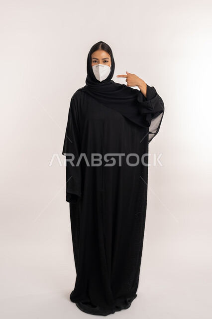 تجنب انتشار العدوى من كوفيد 19، الحفاظ على السلامة, بورتريه لامرأة عربية خليجية إماراتية محجبة ترتدي العباءة السوداء تتبع الإجراءات الاحترازية للوقاية من فايروس كورونا, شابة سعودية تشير بإصبع السبابة إلى القناع الطبي الوقائي, صورة للجسم بالطول الكامل, خلفية رمادية