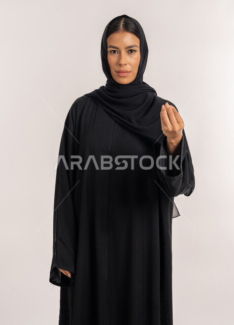 Giving some time to do something, expressions asking to wait, portrait of a veiled Arab Gulf Emirati woman wearing a black abaya and looking at the camera, a young Saudi woman pointing with her hand with gestures of patience and patience, gray background