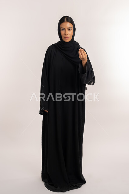 Expressions of asking to wait, giving some time to do something, portrait of a veiled Arab Gulf Emirati woman wearing a black abaya and looking at the camera, a young Saudi woman pointing with her hand with gestures of patience and patience, full-length body portrait, gray background