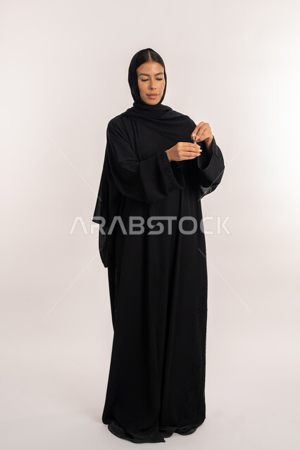 التسويق لمنتجات العطور النسائية, تولة زجاجية من زيت المسك الإماراتي الأصلي, بورتريه لامرأة عربية خليجية إماراتية محجبة ترتدي العباءة السوداء تقوم بالتعطر والتطيب بالزيوت الطبيعية, شابة سعودية تقوم بوضع العطر على يدها, صورة للجسم بالطول الكامل, خلفية رمادية