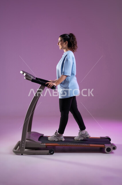 اهمية التمرين لبناء جسد قوي، بورتريه من الجانب لشابة عربية سعودية خليجية مبتسمة ترتدي الملابس الرياضية وتضع سماعة الهيدفون تقوم بالتدريب على جهاز المشي بحماس، المحافظة على نظام الحياة الصحي, أنشطة شبابية تساعد على اللياقة البدنية, صورة الجسم بالطول الكامل, خلفية بنفسجية