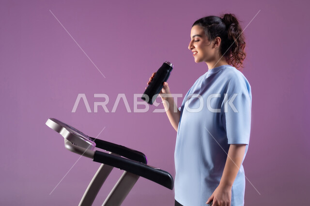 أنشطة رياضية شبابية تساعد على اللياقة البدنية, جودة الحياة الصحية، بورتريه مقرب لشابة عربية خليجية سعودية ترتدي الملابس الرياضية تقوم بشرب الماء أثناء التدريب على العجلة الهوائية، دراجة تمارين ثابتة، خلفية بنفسجية