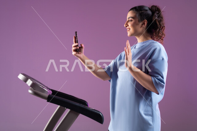 The use of modern technical devices, the concept of agility and fitness, a healthy lifestyle, a close-up portrait of a Saudi Arabian Gulf woman wearing sports clothing, putting on the headphone and making a video call via mobile phone, saying hello and greetings, having a good time while exercising, purple background