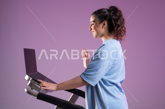 استخدام الأجهزة التقنية الحديثة، مفهوم الرشاقة واللياقة، أسلوب حياة صحية، بورتريه مقرب لإمرأة عربية سعودية خليجية ترتدي الملابس الرياضية وتجري مكالمة فيديو عبر  اللابتوب، القاء التحية والسلام، قضاء وقت ممتع أثناء التمرين، خلفية بنفسجية
