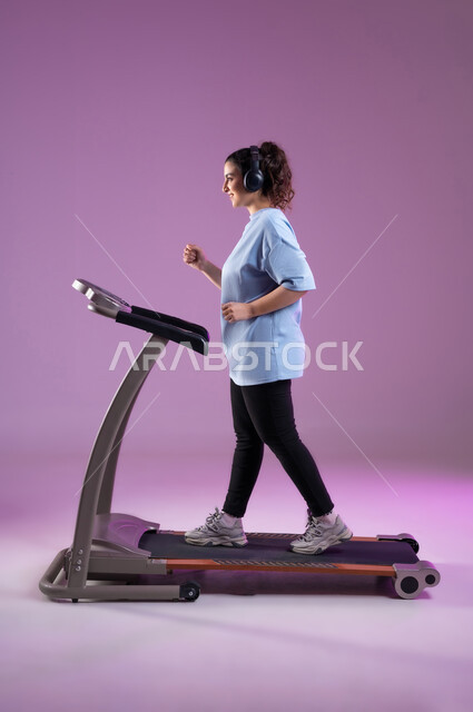 أنشطة شبابية تساعد على اللياقة البدنية, اهمية التمرين لبناء جسد قوي، بورتريه من الجانب لشابة عربية سعودية خليجية مبتسمة ترتدي الملابس الرياضية وتضع سماعة الهيدفون تقوم بالتدريب على جهاز المشي بحماس، المحافظة على نظام الحياة الصحي, صورة الجسم بالطول الكامل, خلفية بنفسجية