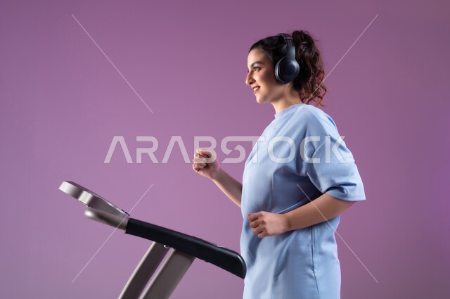 اهمية التمرين لبناء جسد قوي، بورتريه مقرب من الجانب لشابة عربية سعودية خليجية مبتسمة ترتدي الملابس الرياضية وتضع سماعة الهيدفون تقوم بالتدريب على جهاز المشي بحماس، المحافظة على نظام الحياة الصحي, أنشطة شبابية تساعد على اللياقة البدنية, خلفية بنفسجية