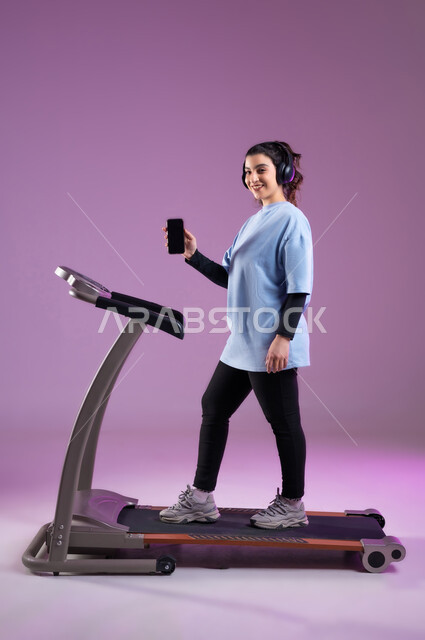 ممارسة الهوايات المفيدة، عرض شاشة سوداء فارغة على الهاتف المحمول، قضاء وقت ممتع على العجلة الهوائية، دراجة تمارين ثابتة، بورتريه لفتاة عربية سعودية خليجية ترتدي الملابس الرياضية وتضع الهيدفون، المحافظة على نظام الحياة الصحي، الطول الكامل للجسم، خلفية بنفسجية