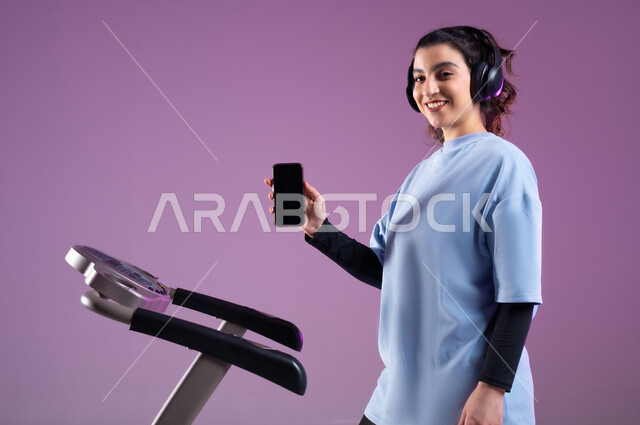 Practicing useful hobbies, displaying a blank black screen on the mobile phone, spending fun time on the bicycle, a stationary exercise bike, a portrait of a Saudi Gulf Arab girl wearing sports clothes and wearing a headphone, maintaining a healthy lifestyle, full length of the body, purple background