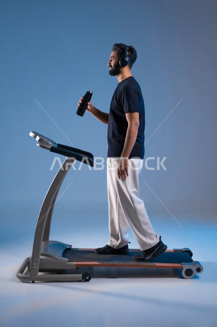 نشاط بدني للمحافظة على اللياقة الجسدية, جودة الحياة الصحية، بورتريه من الجانب لشاب عربي خليجي سعودي مبتسم يرتدي الملابس الرياضية ويضع الهيدفون يقوم بشرب الماء أثناء التدريب على جهاز المشي، الطول الكامل للجسم، خلفية زرقاء