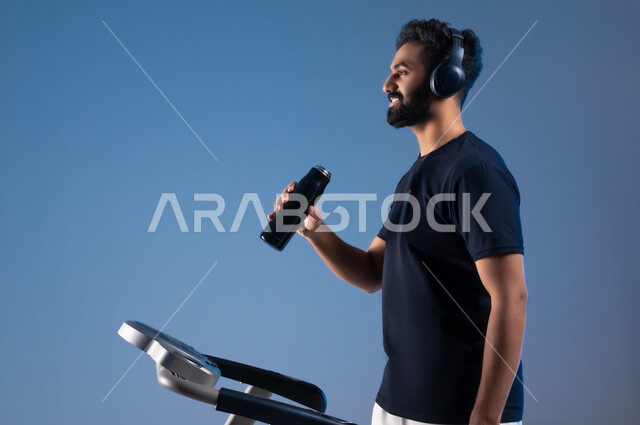 جودة الحياة الصحية, نشاط بدني للمحافظة على اللياقة الجسدية، بورتريه مقرب من الجانب لشاب عربي خليجي سعودي مبتسم يرتدي الملابس الرياضية ويضع الهيدفون يقوم بشرب الماء أثناء التدريب على جهاز المشي، خلفية زرقاء