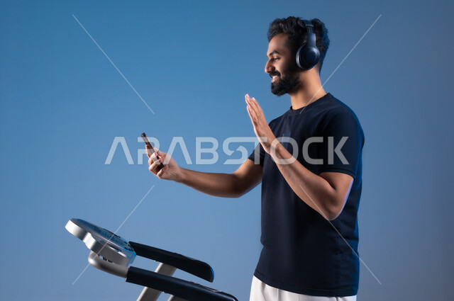 مفهوم الرشاقة واللياقة، استخدام الأجهزة التقنية الحديثة،  أسلوب حياة صحية، بورتريه مقرب من الجانب لشاب عربي سعودي خليجي يرتدي الملابس الرياضية يضع الهيدفون ويقوم بإجراء مكالمة فيديوعبر الهاتف المحمول،القاء التحية والسلام، قضاء وقت ممتع أثناء التمرين،خلفية زرقاء