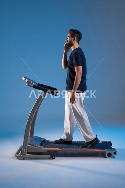 قضاء وقت ممتع أثناء التمرين, مفهوم الرشاقة واللياقة، استخدام الأجهزة التقنية الحديثة، أسلوب حياة صحية، بورتريه من الجانب لشاب عربي سعودي خليجي يرتدي الملابس الرياضية يضع الهيدفون ويقوم بإجراء مكالمة عبر الهاتف المحمول, صورة الجسم بالطول الكامل، خلفية زرقاء