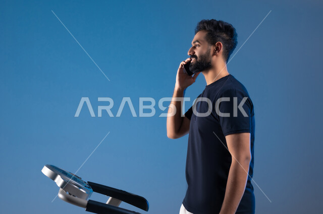 مفهوم الرشاقة واللياقة، استخدام الأجهزة التقنية الحديثة، أسلوب حياة صحية، بورتريه مقرب من الجانب لشاب عربي سعودي خليجي يرتدي الملابس الرياضية يضع الهيدفون ويقوم بإجراء مكالمة عبر الهاتف المحمول، قضاء وقت ممتع أثناء التمرين،خلفية زرقاء