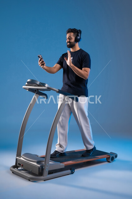 The use of modern technical devices, the concept of agility and fitness, a healthy lifestyle, a close-up portrait of a Saudi Gulf Arab young man wearing sports clothing, putting the headphone on his head and making a video call via mobile phone, saying hello and saying hello, having a good time while exercising, blue background