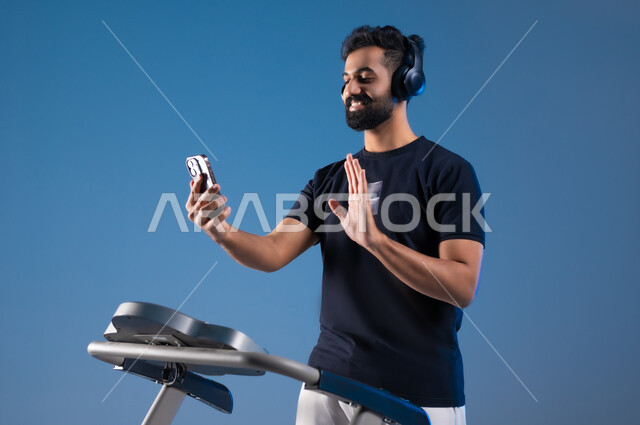 استخدام الأجهزة التقنية الحديثة، مفهوم الرشاقة واللياقة، أسلوب حياة صحية، بورتريه مقرب لشاب عربي سعودي خليجي يرتدي الملابس الرياضية يضع الهيدفون على رأسه ويقوم بإجراء مكالمة فيديوعبر الهاتف المحمول،القاء التحية والسلام، قضاء وقت ممتع أثناء التمرين،خلفية زرقاء