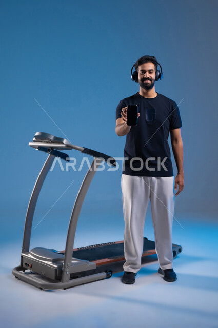 ممارسة الهوايات المفيدة، عرض شاشة سوداء فارغة على الهاتف المحمول، قضاء وقت ممتع على العجلة الهوائية، دراجة تمارين ثابتة، بورتريه لشااب عربي سعودي خليجي يرتدي الملابس الرياضية ويضع الهيدفون، المحافظة على نظام الحياة الصحي، الطول الكامل للجسم، خلفية زرقاء