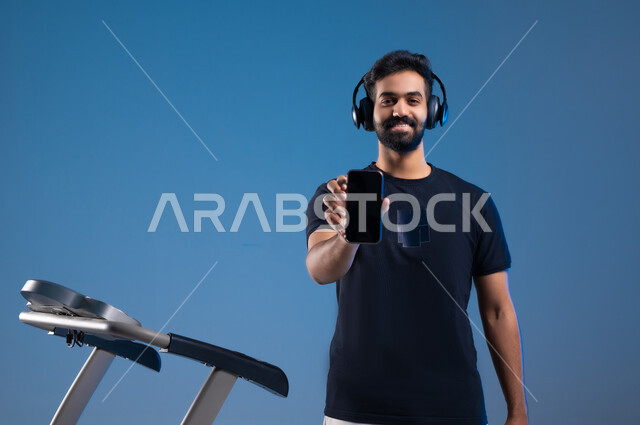 قضاء وقت ممتع على العجلة الهوائية، دراجة تمارين ثابتة، بورتريه مقرب لشاب عربي سعودي خليجي يرتدي الملابس الرياضية ويضع الهيدفون، ممارسة الهوايات المفيدة، عرض شاشة سوداء فارغة على الهاتف المحمول، المحافظة على نظام الحياة الصحي، خلفية زرقاء