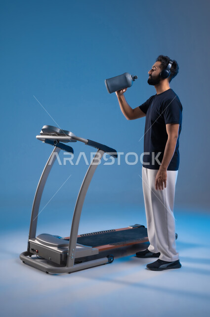 نشاط بدني للمحافظة على اللياقة الجسدية, جودة الحياة الصحية، بورتريه من الجانب لشاب عربي خليجي سعودي مبتسم يرتدي الملابس الرياضية ويضع الهيدفون يقوم بشرب الماء أثناء التدريب على جهاز المشي, صورة الجسم بالطول الكامل، خلفية زرقاء