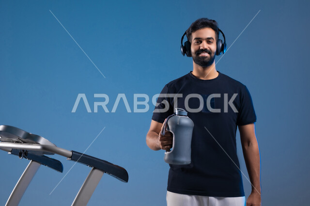 جودة الحياة الصحية، نشاط بدني للمحافظة على اللياقة الجسدية، بورتريه مقرب لشاب عربي خليجي سعودي مبتسم يرتدي الملابس الرياضية ويضع الهيدفون يقوم بشرب الماء أثناء التدريب على جهاز المشي، خلفية زرقاء