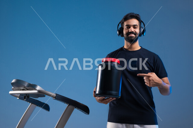 ممارسة الهوايات المفضلة، بورتريه مقرب لشاب عربي خليجي سعودي يرتدي يرتدي ملابس الرياضة ويضع على رأسه سماعة الهيدفون يشير باصبعه السبابة على علبة المكمل الغذائي، المحافة على الرشاقة واللياقة البدنية، مفهوم حرق الدهون وانقاص الوزن خلفية زرقاء
