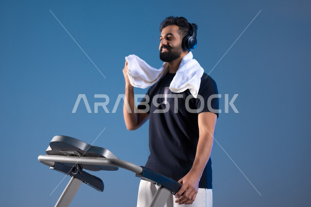 التدرب بعزيمة للمحافظة على نظام الحياة الصحي، انشطة شبابية تساعد على اللياقة البدنية, اهمية الرياضة لبناء جسد قوي, بورتريه مقرب لشاب عربي خليجي سعودي ترتدي الملابس الرياضية ويضع سماعة الهيدفون يستخدم المنشفة لتجفيف العرق أثناء التمرين، خلفية زرقاء