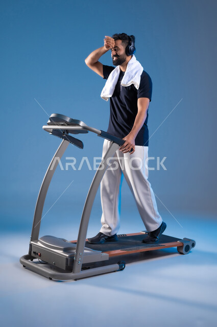 Feeling tired and exhausted during exercise, training with determination to maintain a healthy lifestyle, youth activities that help with physical fitness, the importance of sports to build a strong body, a close-up portrait of a young Saudi Arabian Gulf man wearing sports clothes and wearing a headphone, full-length body portrait, blue background