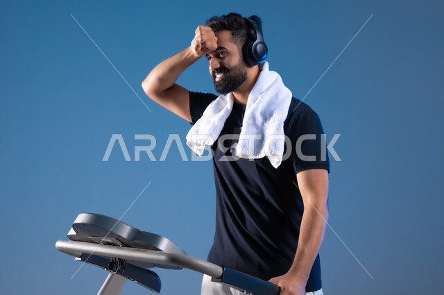 الشعور بالتعب والارهاق اثناء التمرين, التدرب بعزيمة للمحافظة على نظام الحياة الصحي، انشطة شبابية تساعد على اللياقة البدنية, اهمية الرياضة لبناء جسد قوي, بورتريه مقرب لشاب عربي خليجي سعودي يرتدي الملابس الرياضية ويضع سماعة الهيدفون ، خلفية زرقاء