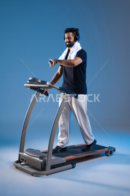 Maintaining a healthy lifestyle, gestures of happiness and activity, the concept of agility and fitness, a portrait of a Saudi Gulf Arab young man wearing sports clothing, placing a headphone on his head, following his progress in performance on a treadmill, practicing useful hobbies, full body length, blue background.