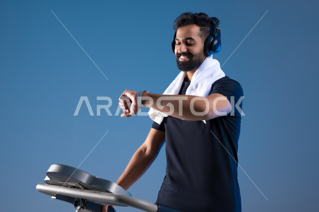 التدرب بعزيمة للمحافظة على نظام الحياة الصحي، ايماءات السعادة والنشاط، مفهوم الرشاقة واللياقة، بورتريه مقرب لشاب عربي خليجي سعودي يرتدي الملابس الرياضية يضع الهيدفون على رأسه يتابع تقدمه في الأداء على جهاز المشي، ممارسة الهوايات المفيدة، خلفية زرقاء