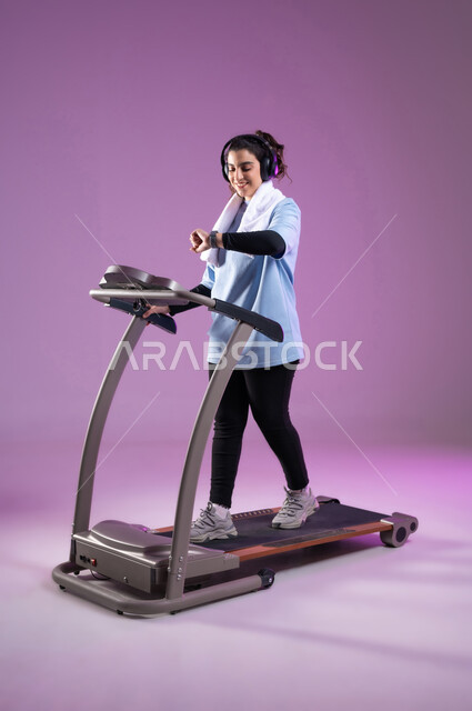 ممارسة الهوايات المفيدة، ايماءات السعادة والنشاط، مفهوم الرشاقة واللياقة، بورتريه لفتاة عربية خليجية سعودية ترتدي الملابس الرياضية تضع الهيدفون وتتابع تقدمها في الأداء على جهاز المشي، التدرب بعزيمة للمحافظة على نظام الحياة الصحي،الطول الكامل للجسم خلفية بنفسجية