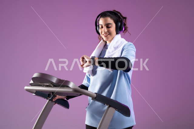ايماءات السعادة والنشاط، مفهوم الرشاقة واللياقة، بورتريه مقرب لفتاة عربية خليجية سعودية ترتدي الملابس الرياضية تضع الهيدفون على رأسها تتتابع تقدمها في الأداء على جهاز المشي، ممارسة الهوايات المفيدة، التدرب بعزيمة للمحافظة على نظام الحياة الصحي، خلفية بنفسجية