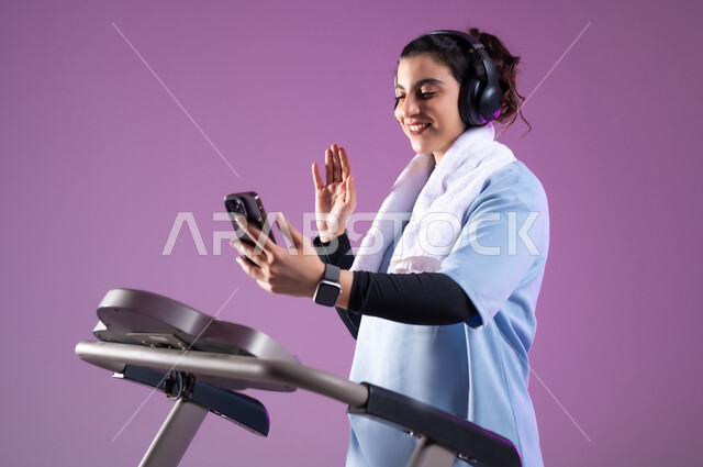 القاء التحية والسلام، استخدام الأجهزة التقنية الحديثة، مفهوم الرشاقة واللياقة، أسلوب حياة صحية، بورتريه مقرب لإمرأة عربية سعودية خليجية ترتدي الملابس الرياضية تضع الهيدفون وتجري مكالمة فيديوعبر الهاتف المحمول، قضاء وقت ممتع أثناء التمرين، خلفية بنفسجية